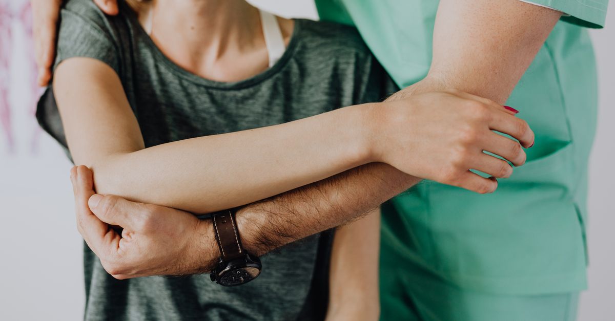 Crop anonymous chiropractor in uniform and wristwatch examining shoulder of smiling faceless woman in casual clothes in doctor office in clinic